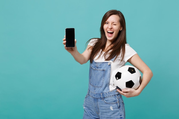 Una alegre fanática del fútbol que parpadea sosteniendo una pelota de fútbol, un teléfono móvil con una pantalla vacía en blanco aislada en un fondo azul turquesa. Emociones de la gente, concepto de ocio familiar deportivo. Simulacros de espacio de copia.