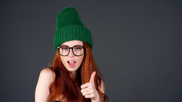 Alegre feliz mulher jovem e bonita com cabelo vermelho, com um chapéu de malha verde mostra um polegar para