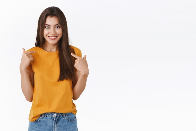 Alegre, feliz, triunfante, joven mujer bonita, afortunada ganadora del premio o fue elegida para la promoción, señalándose orgullosa, fanfarroneando, logro personal, sonriendo alegremente, fondo blanco.