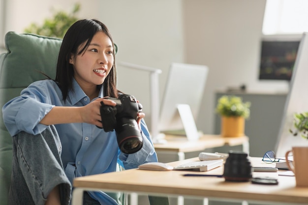 Alegre fotógrafa vietnamita segurando a câmera fotográfica sentada no local de trabalho