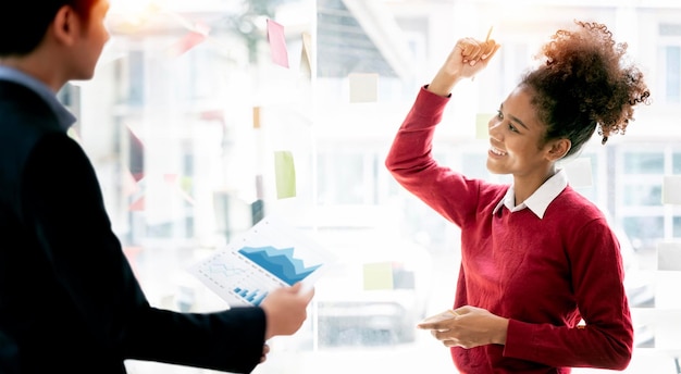 Alegre freelancer étnico positivo olhando para o post-it na parede e segurando os braços para cima gritando comemorando a conquista da vitória projeto concluído com sucesso