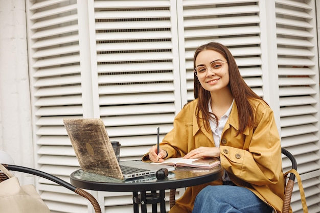 Alegre freelancer feminino trabalhando no café de rua e olhando para a câmera enquanto escreve no caderno