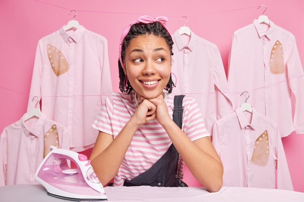 Foto alegre garota afro-americana com dreadlocks na tábua de passar sorrisos desvia o olhar, afasta as roupas ocupadas fazendo trabalhos domésticos na lavanderia usa t-shirt com tiara e macacão.