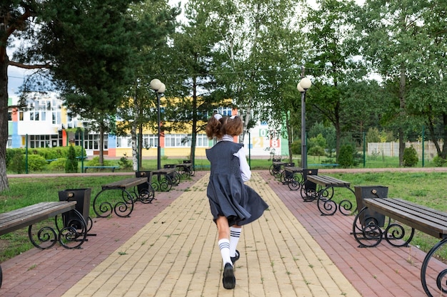 Alegre garota engraçada com sorriso desdentado em uniforme escolar com arcos brancos correndo no pátio da escola De volta à escola 1º de setembro Aluno feliz com mochila Movimento de classe primária de educação primária