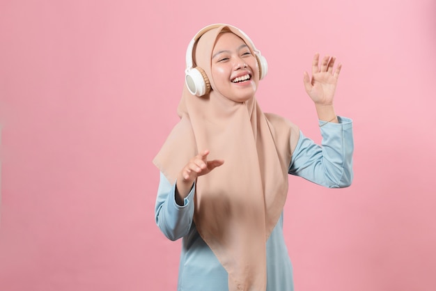 Alegre garota muçulmana asiática ouvindo música em fones de ouvido sem fio e dançando sobre fundo rosa