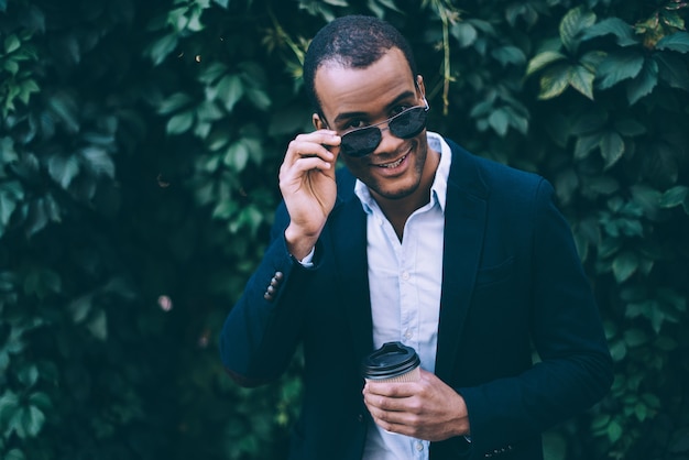 Alegre guapo. Apuesto joven africano ajustando sus gafas de sol y sonriendo mientras sostiene la taza de café y de pie contra el fondo de la planta verde al aire libre