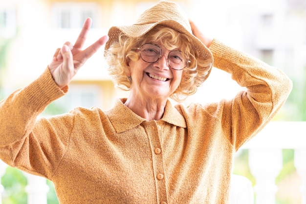 Alegre hermosa anciana en amarillo con sombrero y anteojos mirando a la cámara haciendo gestos de victoria