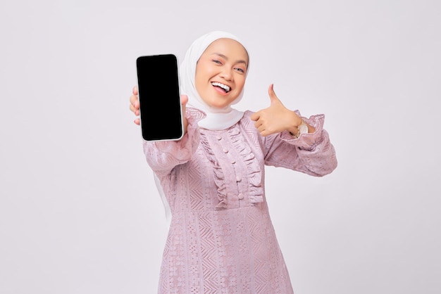 Una alegre y hermosa joven musulmana asiática con hiyab y vestido morado demostrando un smartphone con una pantalla vacía que muestra un gesto con el pulgar hacia arriba aislado en el fondo del estudio blanco