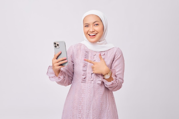 Una alegre y hermosa joven musulmana asiática con hiyab y vestido morado señalando con el dedo al teléfono móvil y recomienda una aplicación aislada en el fondo de un estudio blanco