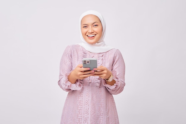 Una alegre y hermosa joven musulmana asiática con hiyab y vestido morado sosteniendo un teléfono móvil y mirando la cámara con confianza aislada en el fondo de un estudio blanco