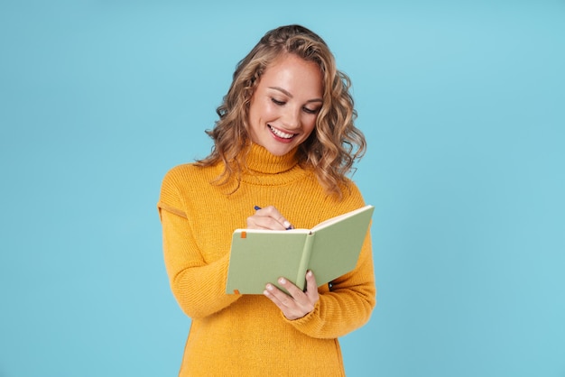 Alegre hermosa joven vistiendo suéter que se encuentran aisladas en azul, escribiendo en un diario