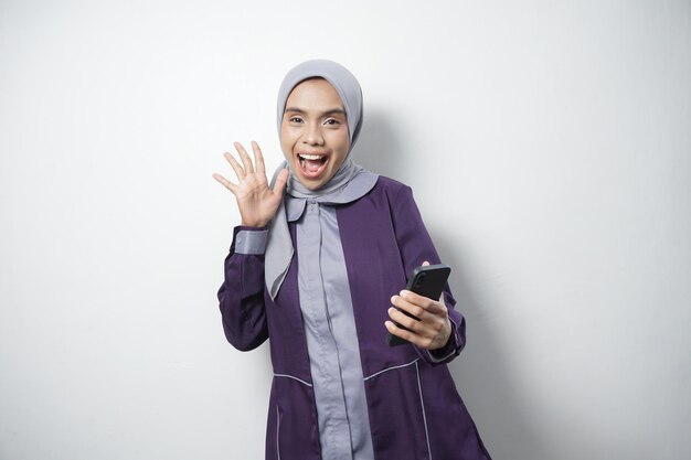 Alegre y hermosa mujer asiática con camisa casual y hijab usando un teléfono móvil aislado por un fondo blanco