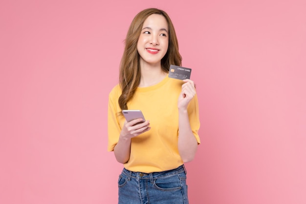 Alegre hermosa mujer asiática con smartphone y tarjeta de crédito de maqueta sobre fondo rosa