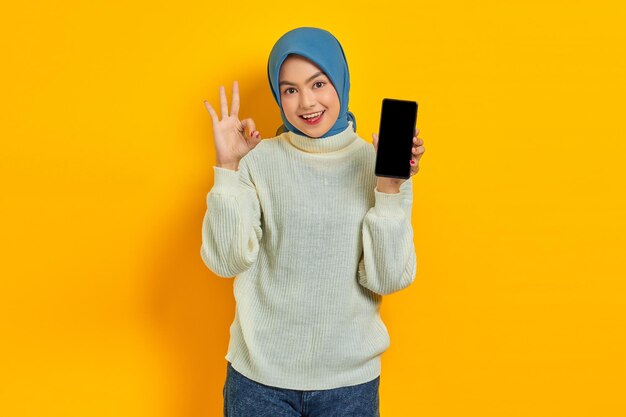 Alegre hermosa mujer asiática con suéter blanco y hiyab que muestra una pantalla de teléfono móvil en blanco que muestra un gesto correcto aislado en un concepto de estilo de vida musulmán de fondo amarillo