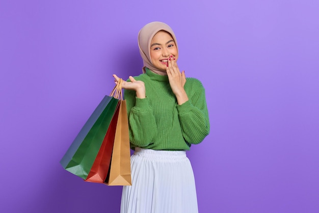 Alegre hermosa mujer asiática en suéter verde sosteniendo bolsas de la compra con la mano en la boca aislada sobre fondo púrpura