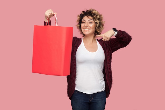 Foto alegre hermosa mujer joven en camisa azul con peinado rizado de pie y apuntando con el dedo a las bolsas de la compra y una gran sonrisa, mirando a la cámara. foto de estudio, fondo rosa, aislado, interior