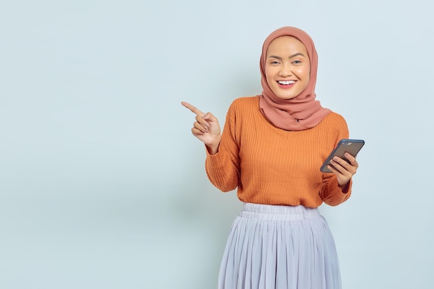 Alegre y hermosa mujer musulmana asiática con suéter marrón e hiyab sosteniendo un teléfono inteligente y señalando con el dedo el espacio de la copia aislado en el concepto de estilo de vida musulmán de fondo blanco