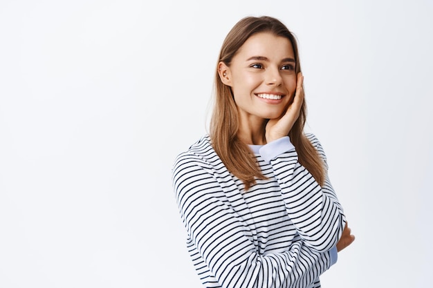 Alegre hermosa mujer sincera sonriendo, mirando el espacio de la copia del lado derecho, tocando la mejilla, la piel sana y natural y los dientes blancos, estudio