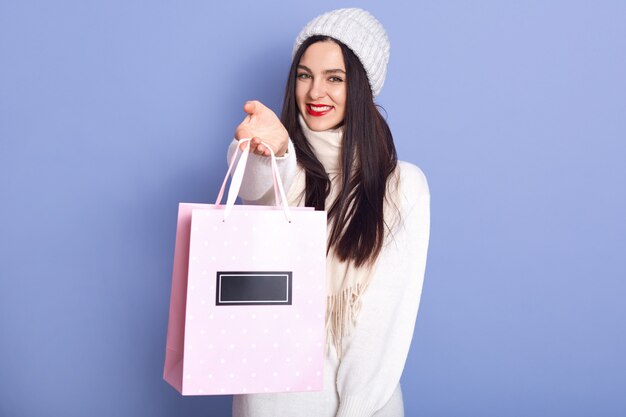 Alegre hermosa mujer vistiendo suéter blanco, sombrero y bufanda, sosteniendo una bolsa de papel