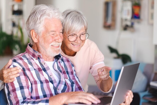 Alegre y hermosa pareja de ancianos sonriendo en casa navegando juntos en una laptop Pareja caucásica activa de ancianos disfrutando de la tecnología y las redes sociales