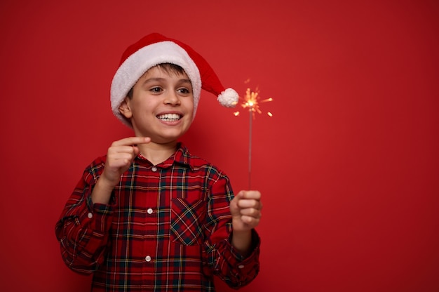 Alegre, hermoso, asombroso, niño, preadolescente, con, sombrero de santa, y, tela escocesa, rojo, camisa, regocijo, mirar, bengala, luces de navidad, bengala, en, el suyo, manos, contra, fondo coloreado, con, espacio de copia, para, anuncio