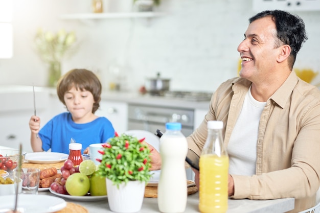 Alegre hispano de mediana edad sonriendo, mientras desayuna con su familia, sentado en la mesa de la cocina cerca de su hijo en casa. Familia, paternidad, concepto de mañana