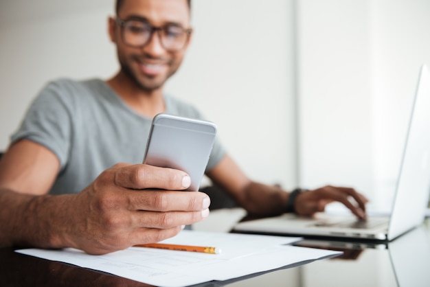 Alegre hombre africano vestido con camiseta gris y gafas usando teléfono celular y sentado en la mesa. Mirando el teléfono. Centrarse en la mano con el teléfono.