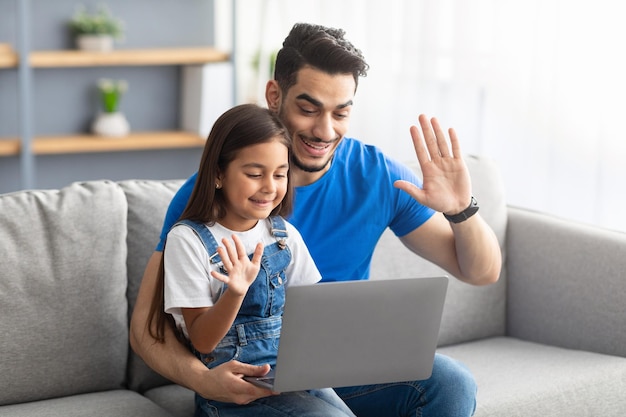 Alegre hombre árabe y su hija haciendo videollamadas usando la computadora portátil en casa, familia feliz teniendo reunión de conferencia en línea con amigos o familiares, saludando con la mano a la cámara, diciendo hola