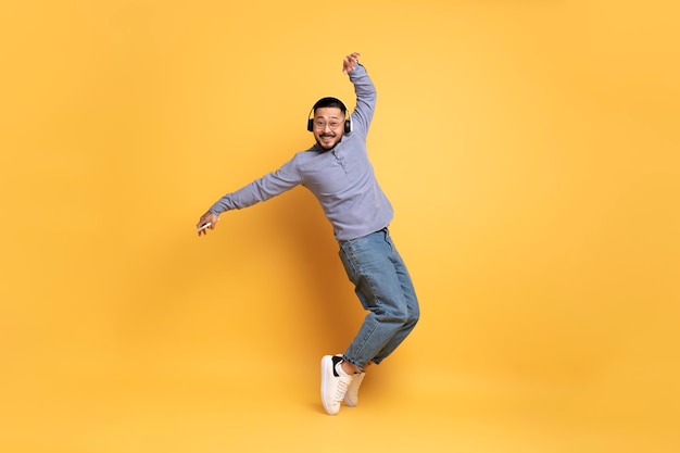 Alegre hombre asiático con auriculares inalámbricos bailando sobre fondo amarillo