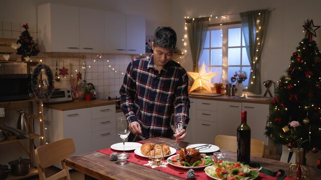alegre hombre asiático poniendo copas de vino mientras prepara la mesa de la cena de Navidad en casa con su pareja femenina cocinando en el fondo en una cocina festiva en casa