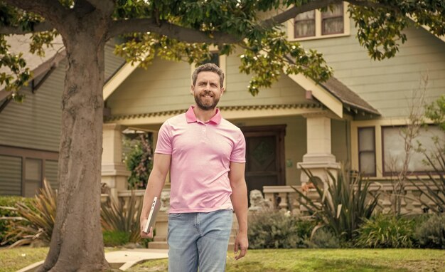 Foto alegre hombre barbudo agente de bienes raíces vendiendo o alquilando casa a pie con vecindario de computadoras