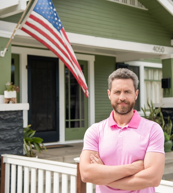 Alegre hombre barbudo agente de bienes raíces vendiendo o alquilando casa con suburbio de bandera americana