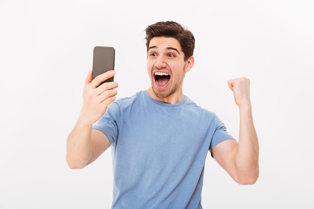 Alegre hombre gritando en camiseta con smartphone y se regocija con los ojos cerrados sobre la pared gris