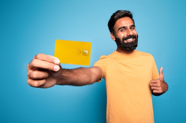 Alegre hombre indio barbudo con tarjeta de crédito mostrando el gesto del pulgar hacia arriba promoviendo el pago sin contacto