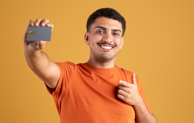 Foto alegre hombre latino mostrando tarjeta de crédito y apuntándola recomendando oferta bancaria de pie en amarillo