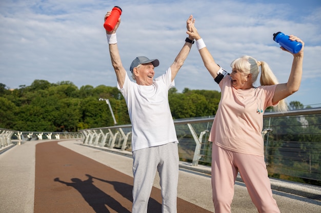 Foto alegre hombre y mujer senior levantan botellas y unen sus manos en la pasarela