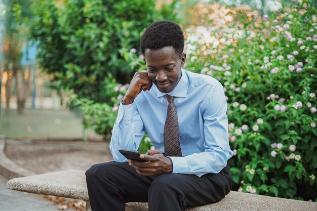 Alegre hombre de negocios africano usando las redes sociales mientras descansa en un banco