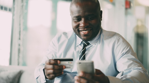 Alegre hombre de negocios afroamericano con ropa formal pagando facturas en línea manteniendo la tarjeta de crédito y el teléfono inteligente en sus manos en un café vidrioso