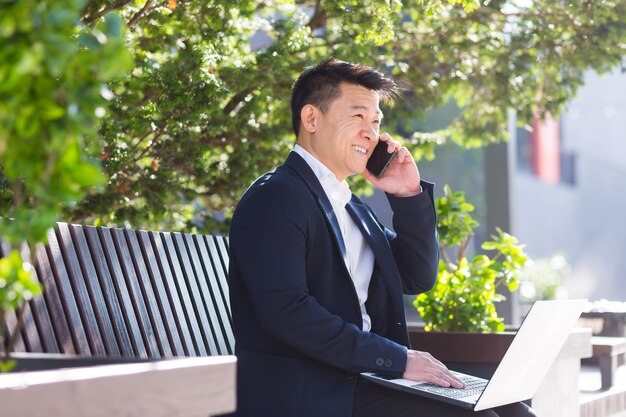 Alegre hombre de negocios asiático trabajando en la computadora portátil y hablando por teléfono, sonriendo sentado en un banco del parque cerca de la oficina en el almuerzo