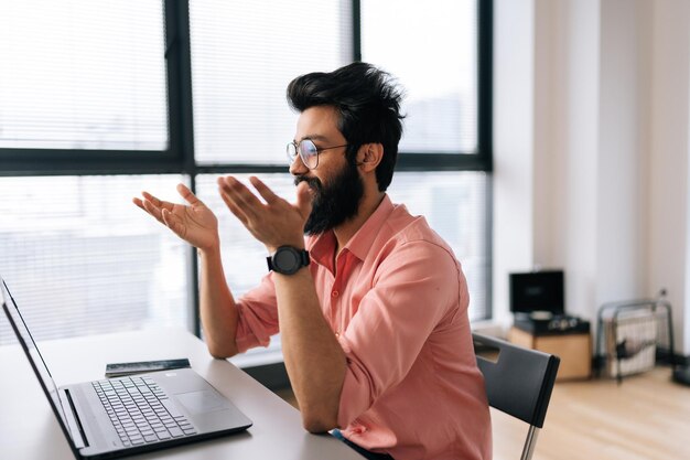Alegre hombre de negocios indio con anteojos hablando en línea por videollamada en una laptop sentada en el escritorio junto a la ventana mirando a la pantalla