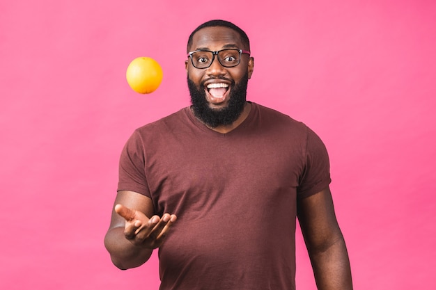 Alegre hombre negro afroamericano sosteniendo una fruta naranja, vitaminas para la salud, concepto de dieta aislado sobre fondo rosa.