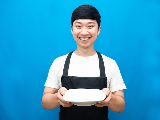 Alegre hombre vestido con delantal dando plato vacío fondo azul.