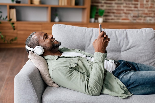 Alegre homem afro-americano maduro em casual em fones de ouvido sem fio gosta de música favorita com smartphone