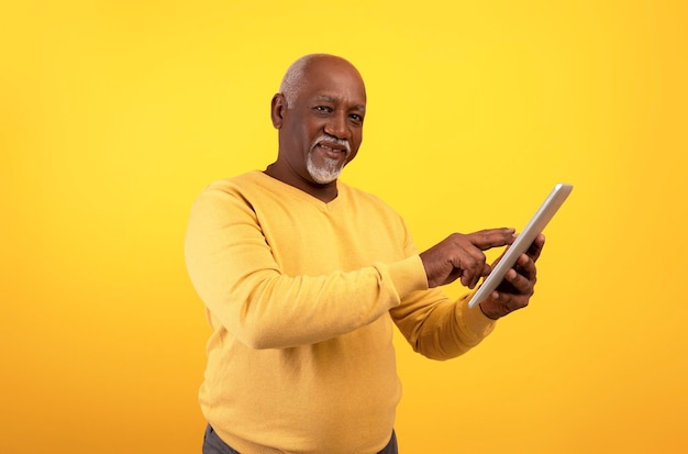 Alegre homem afro-americano sênior usando computador tablet para trabalho on-line ou estudos em fundo de estúdio laranja