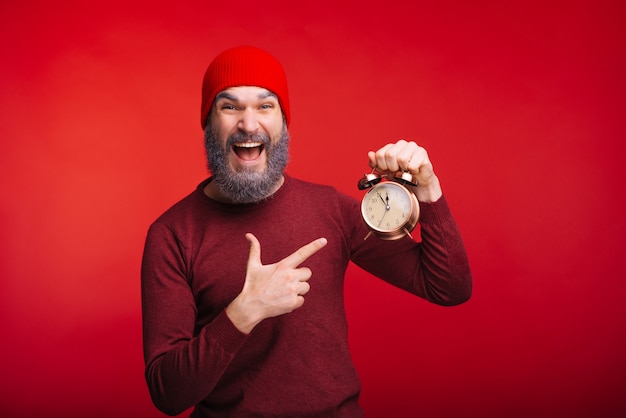 Alegre homem barbudo em cima de espaço vermelho, apontando para o relógio despertador