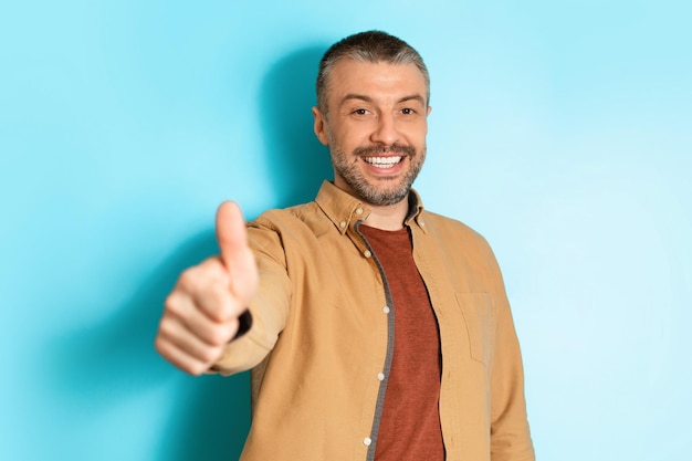 Alegre homem de meia idade gesticulando polegares para cima sobre fundo azul