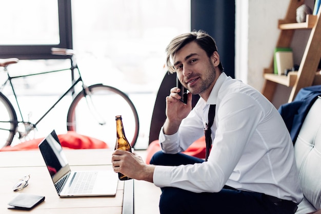 Alegre homem de terno segurando a garrafa e falando no smartphone depois da festa