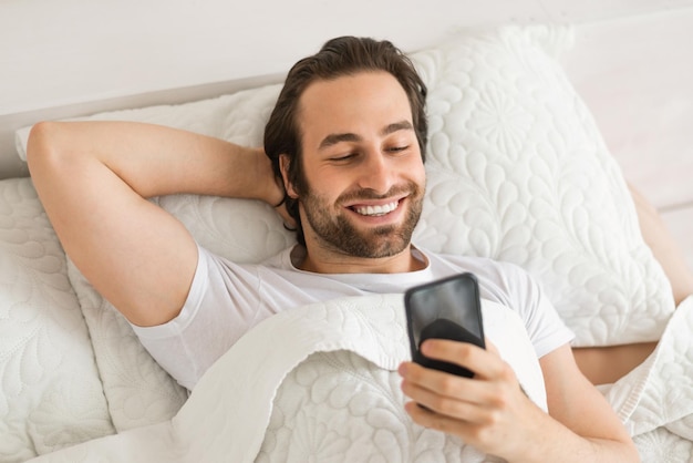 Alegre homem europeu milenar com barba por fazer está na cama branca digitando no smartphone assistindo vídeo