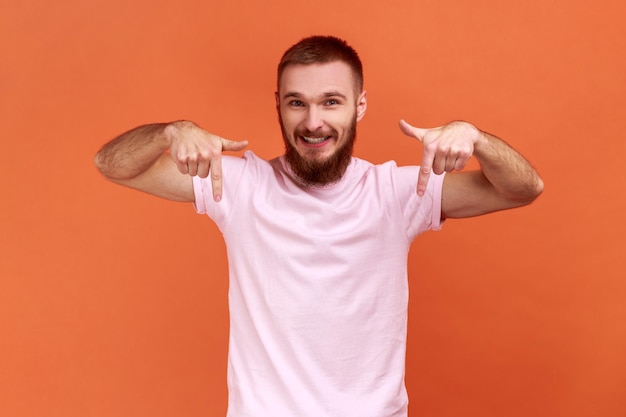 Alegre homem satisfeito apontando os dedos para baixo prestando atenção ao blogueiro da área de publicidade recomendando se inscrever usando camiseta rosa foto de estúdio interior isolada em fundo laranja