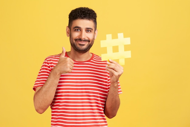 Alegre homem satisfeito com barba em camiseta listrada, mostrando os polegares para cima como gesto segurando o sinal de hashtag amarelo satisfeito com o anúncio alvo Tiro de estúdio interior isolado em fundo amarelo